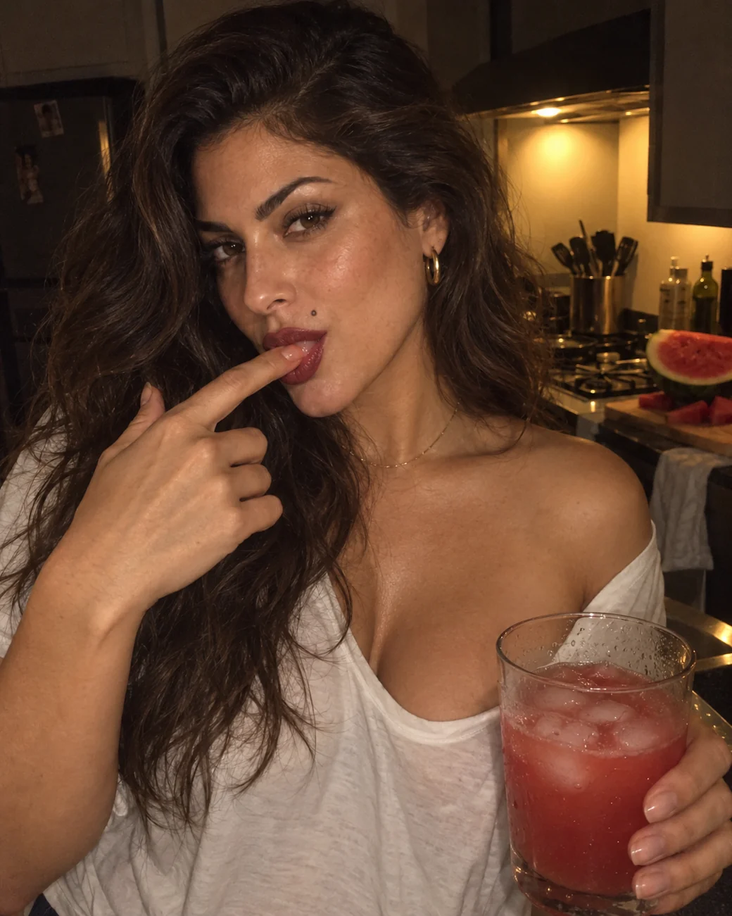 Mulher na cozinha à noite, lambendo o suco do dedo e olhando para a câmera, com um copo de suco de melancia na mão