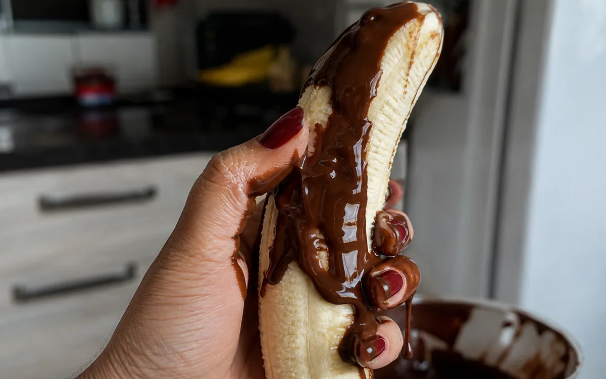 Mão segurando uma banana com calda de chocolate escorrendo